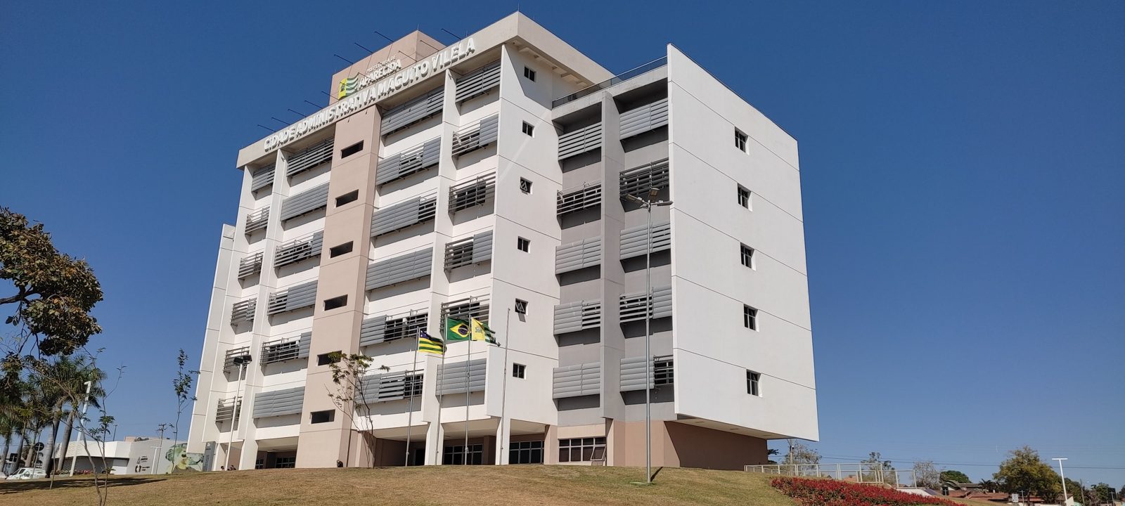 Fachada da Cidade Administrativa Maguito Vilela, sede da Prefeitura de Aparecida de Goiânia, com brises cinza e bandeiras do Brasil, de Goiás e do município